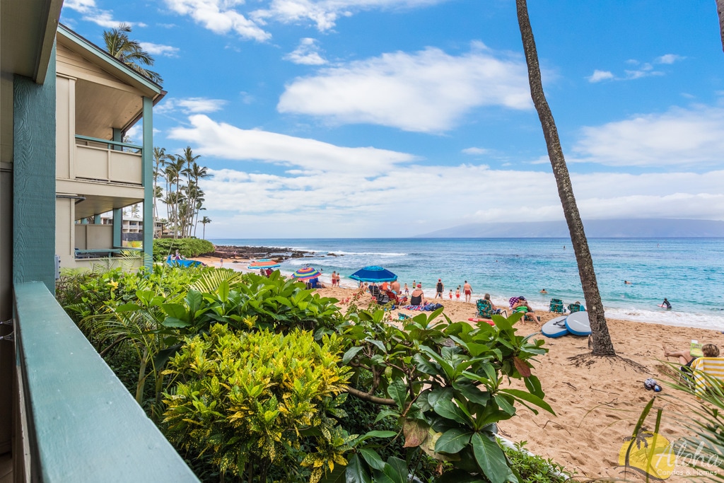 A vibrant view of Napili Bay is showcased, with golden sand and gentle waves meeting the shoreline. Sunbathers and colorful beach umbrellas dot the beach, framed by lush greenery and palm trees. The open sky is filled with soft clouds, enhancing the coastal serenity.