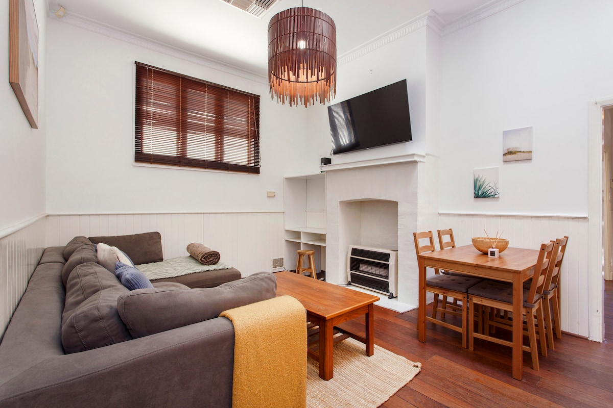 An open-plan living area is presented, featuring a comfortable grey couch with a textured throw, a wooden dining table set for four, and a white fireplace with a television mounted above. Natural light filters through the wooden window blinds, enhancing the inviting space.