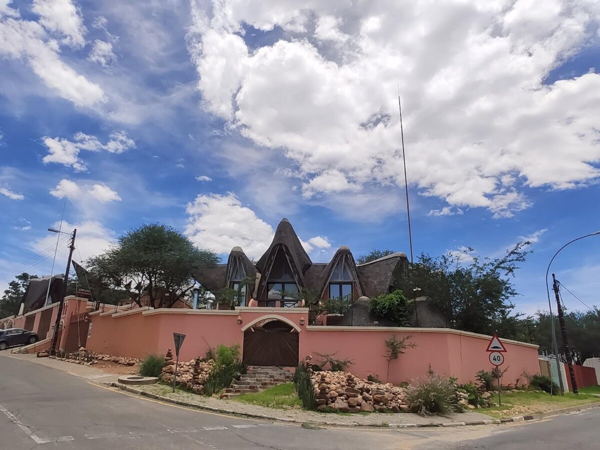 A charming building with a pink exterior is set against a bright sky. Unique thatched roofs adorned with large windows create an inviting appearance. Surrounding greenery and a well-kept garden add to the tranquil atmosphere, while a winding road leads up to the entrance.