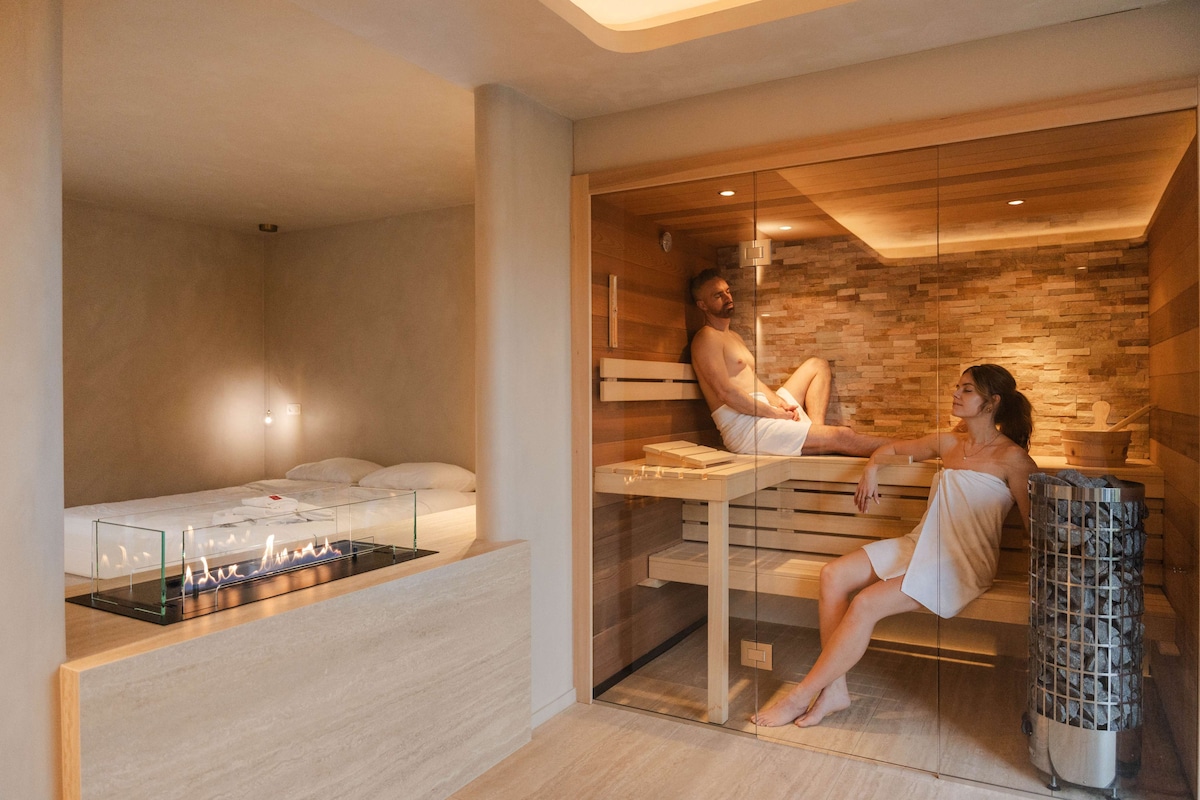 An elegantly designed space features a luxurious kingsize bed adjacent to a modern sauna area. The sauna, with wooden benches and stone accents, is visible behind a glass door. A stylish fireplace adds warmth to the room, creating an intimate atmosphere.