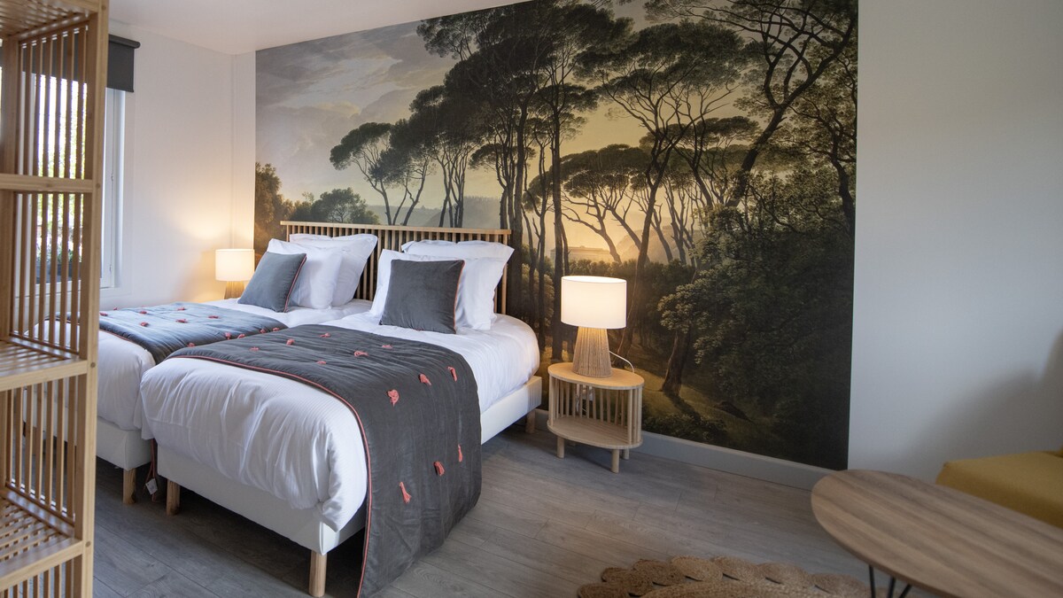 A guest room features two spacious beds dressed with neutral linens and gray accents. A wall mural depicting a serene forest landscape enhances the atmosphere. Soft lighting from two nightstands illuminates the room, while a circular area rug and a small table add warmth to the space.