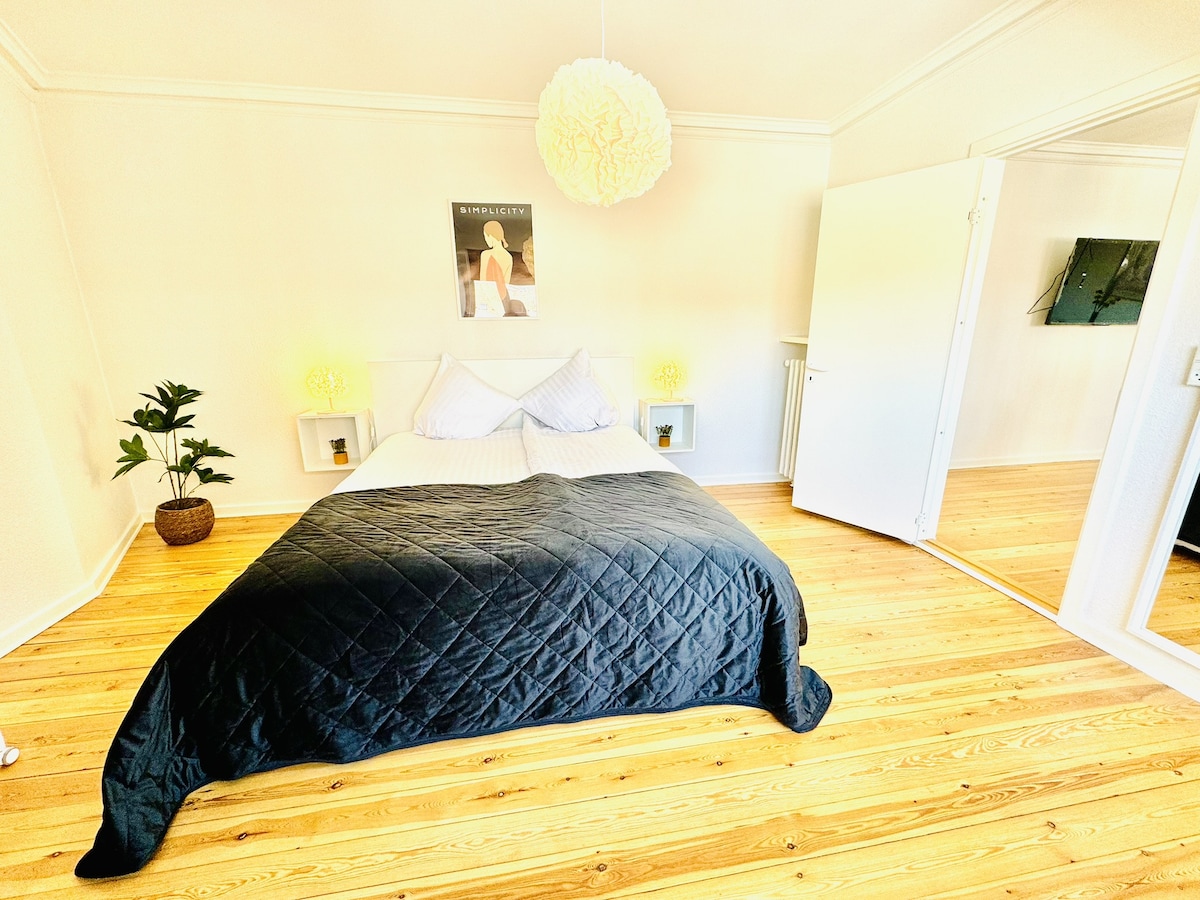A bright bedroom is highlighted by a spacious bed covered with a dark quilt. Soft yellow lighting is provided by bedside lamps. A decorative plant is positioned in the corner, and a wall-mounted television is visible. Mirrored wardrobes reflect the room's openness.