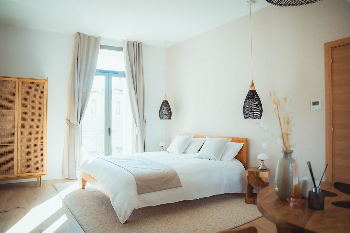 A spacious room features a comfortable bed with neutral bedding and soft pillows. Two woven pendant lights hang above, while natural light pours in through large windows. A wooden wardrobe and a round dining table with decorative elements complete the elegant space.