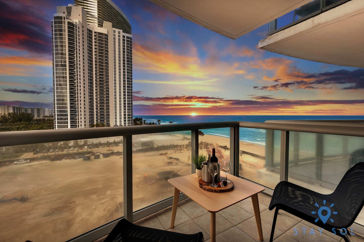 A balcony offers sweeping views of the ocean and the sunset, with vibrant hues of orange and purple across the sky. A small table with two chairs is positioned on the balcony, inviting relaxation and enjoyment of the picturesque scenery.