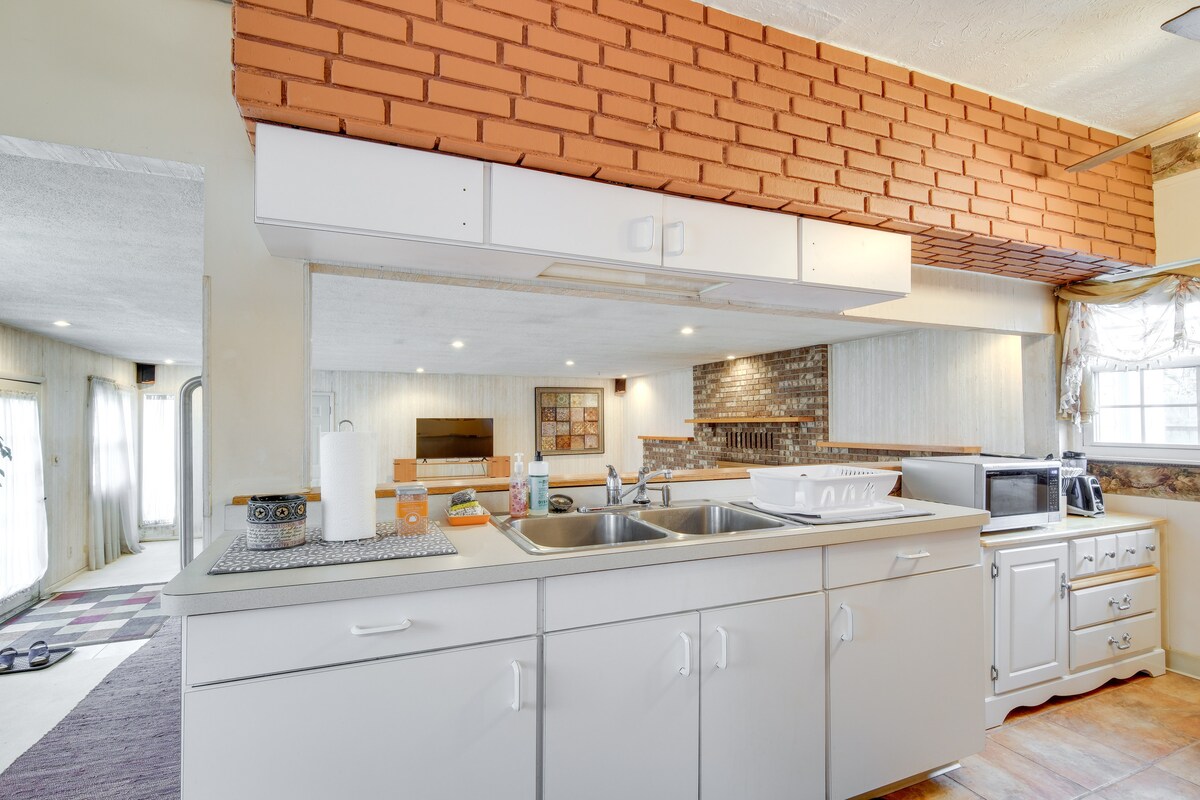 The kitchen area features white cabinetry with ample storage space, alongside a double sink and a drainer. A microwave and stove are visible, with a brick backsplash providing texture. The open layout leads to a spacious living area, enhancing natural light within the space.