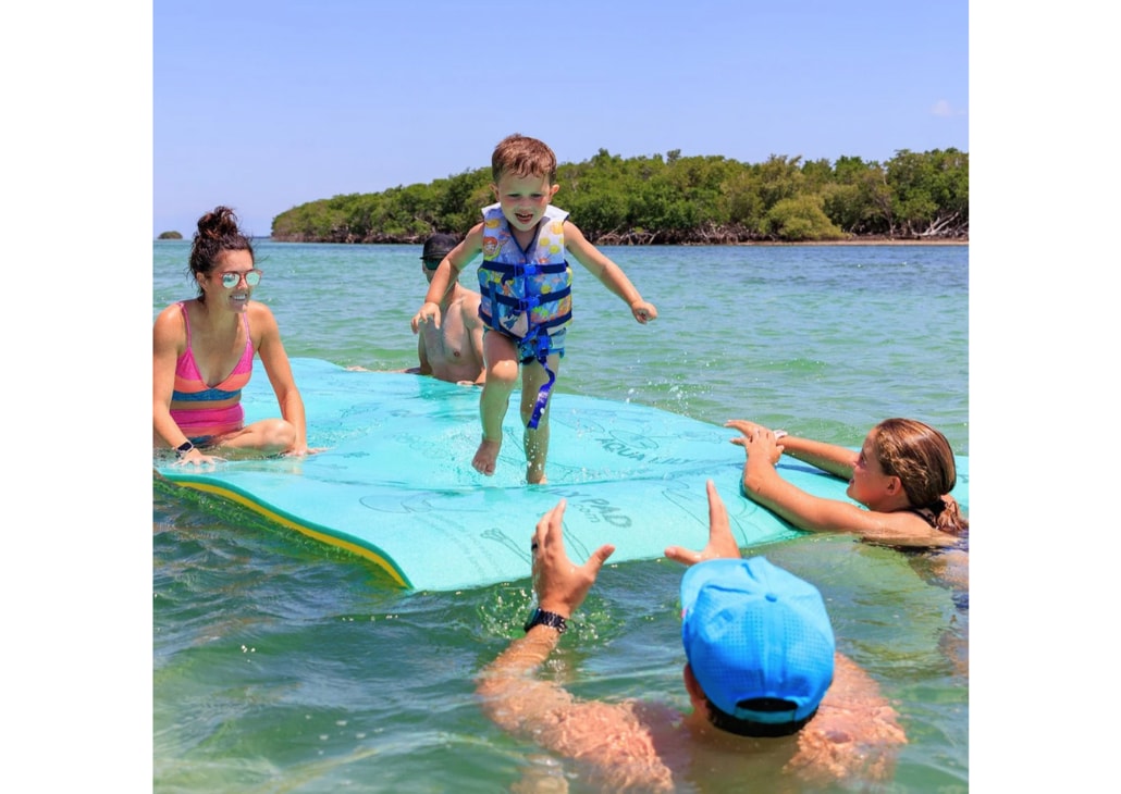 A child is captured mid-air while jumping onto a floating lily pad in a clear body of water. Two adults are positioned in the water, reaching out with welcoming gestures. The surrounding greenery adds a serene, natural backdrop to the scene.