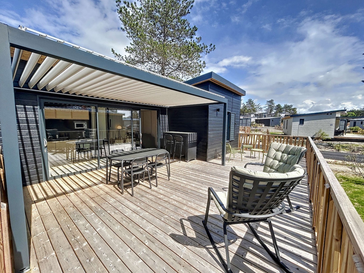 A spacious terrace is shown, featuring a wooden deck that accommodates a dining table and multiple lounge chairs. Sunlight filters through the bioclimatic pergola, creating a comfortable outdoor space. The adjacent modern chalet is visible, alongside additional residence structures in the background.