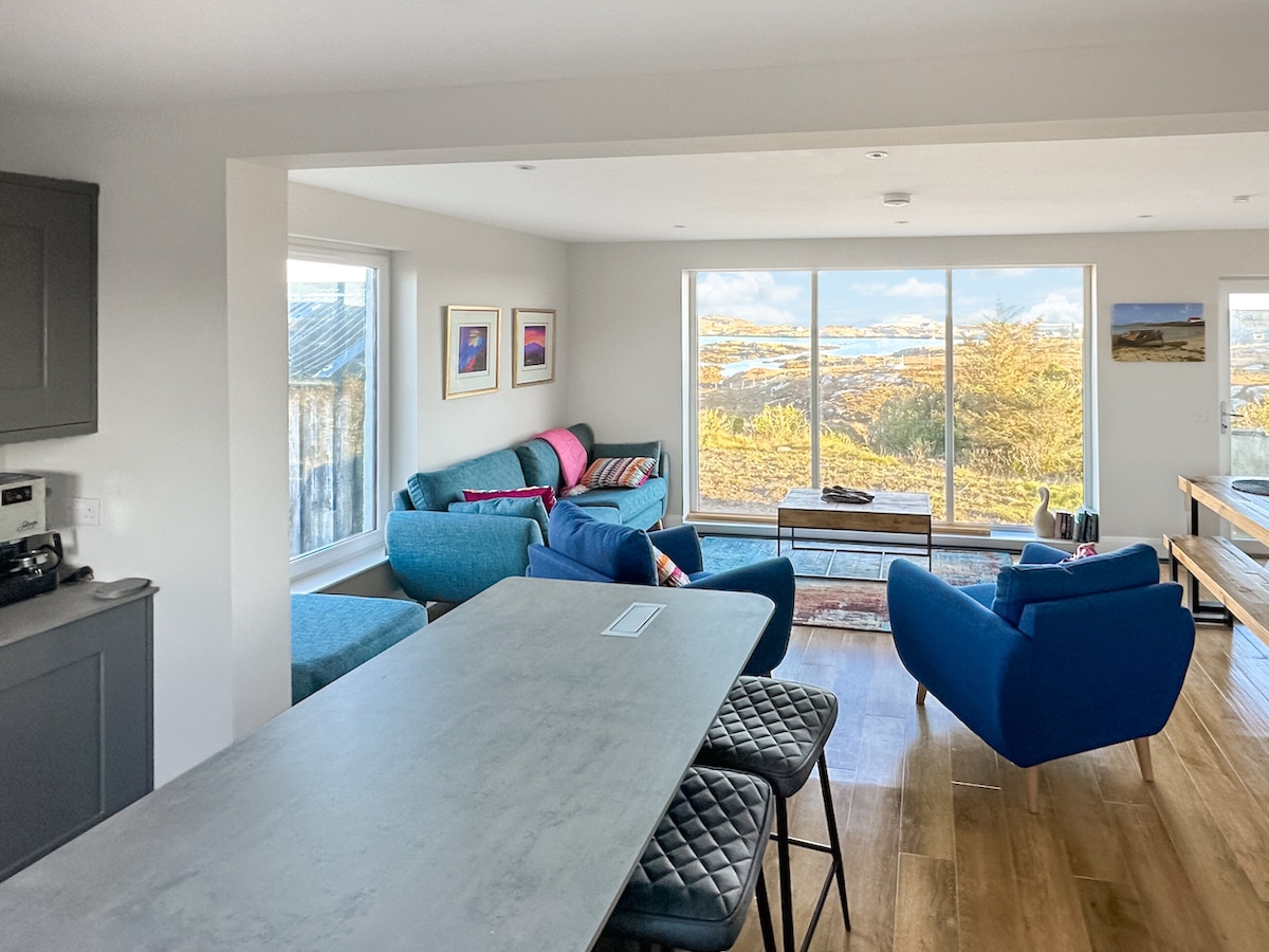 An open plan living area is presented with large windows offering views of the surrounding landscape. Comfortable seating in varying shades is arranged around a central coffee table, complementing a dining area with a substantial grey table. Natural light fills the space, creating a bright and welcoming ambiance.