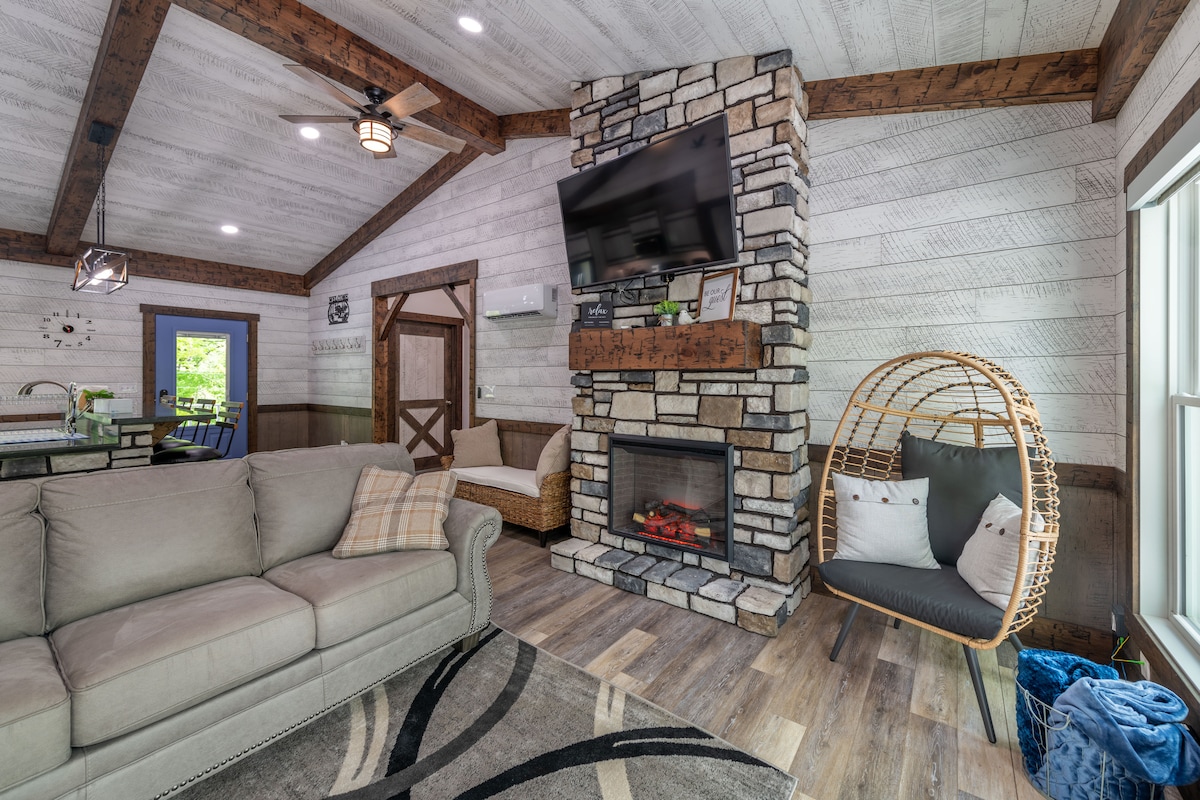 A welcoming living area features a cozy sofa and an elegant stone fireplace with a mounted television above. Natural light fills the room through large windows, and a stylish hanging chair adds a touch of modern comfort. Rustic wooden beams accentuate the ceiling.