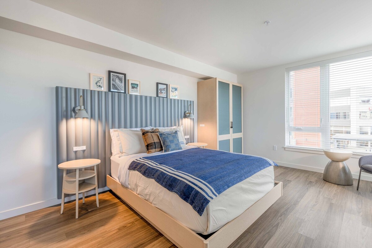 A well-lit bedroom features a spacious bed with a textured blue bedspread and matching pillows. A headboard with vertical slats complements the room's modern design, while framed pictures adorn the wall above. A bedside table and a sculptural side table are included, alongside large windows allowing natural light.