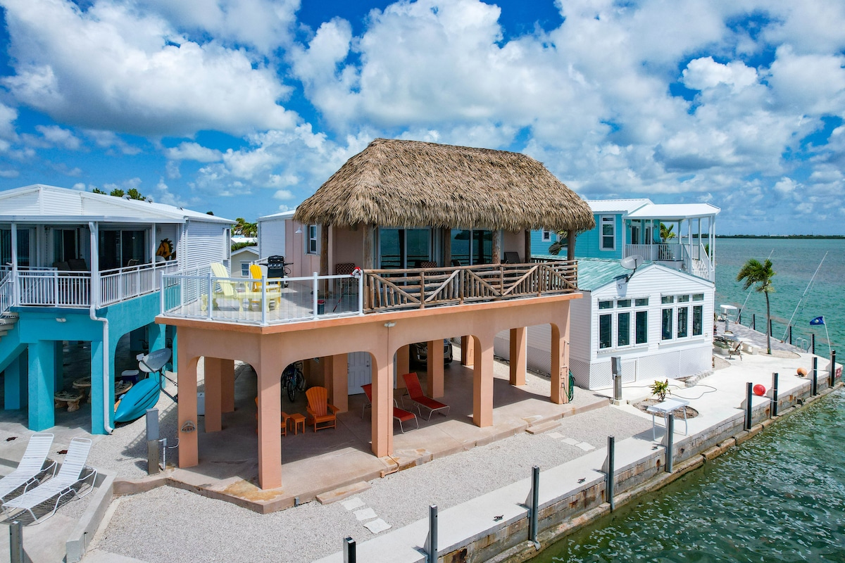 An oceanfront retreat is showcased, featuring a thatched roof and a spacious balcony with seating. The structure is surrounded by calm water and a dock area. Sun loungers are placed along the waterfront, creating areas for relaxation under a bright sky dotted with clouds.