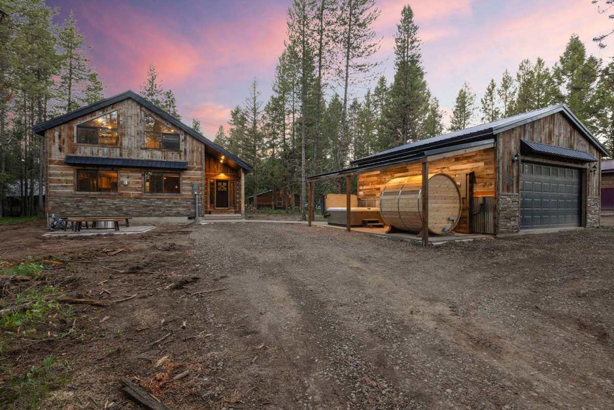 The rustic modern cabin is surrounded by tall trees, featuring a spacious front gravel area. A detached garage is visible, along with a barrel sauna, presenting inviting outdoor amenities. The soft glow of sunset colors the sky, enhancing the tranquil setting.