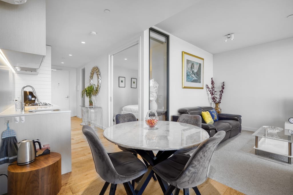 A modern dining area features a round marble table surrounded by plush velvet chairs. In the background, a cozy living space is visible, with a dark sofa and decorative elements such as art and plants. Natural light enhances the open layout and neutral color palette.