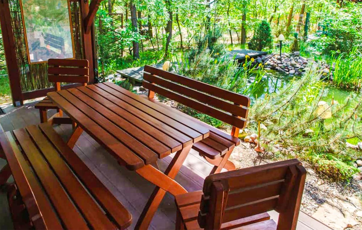 A spacious wooden table and bench seating are positioned under a covered terrace. The surrounding area features lush greenery and a tranquil pond, creating a serene outdoor setting ideal for dining or relaxing. Light filters softly through the trees, enhancing the peaceful atmosphere.