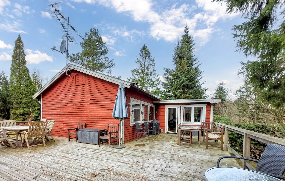 A bright red vacation home is set against a backdrop of trees. A spacious wooden terrace features multiple seating arrangements, including chairs and tables. An umbrella and a satellite dish are also visible, with large glass doors leading into the home.