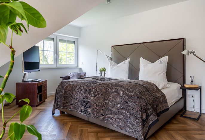 The spacious sleeping area features a plush bed with decorative pillows and a patterned throw blanket. A modern side table and reading lamp are positioned beside the bed. Natural light enters through nearby windows, illuminating a cozy seating area with a chair and a television.