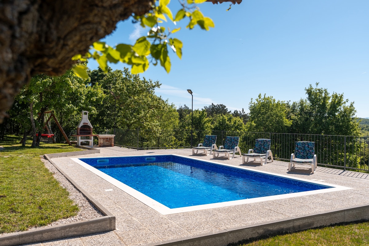 A serene outdoor area features a vibrant blue swimming pool surrounded by a sun terrace, equipped with multiple lounge chairs. Lush greenery provides a natural backdrop, while a barbecue area and children's playground are also visible, enhancing the outdoor experience.