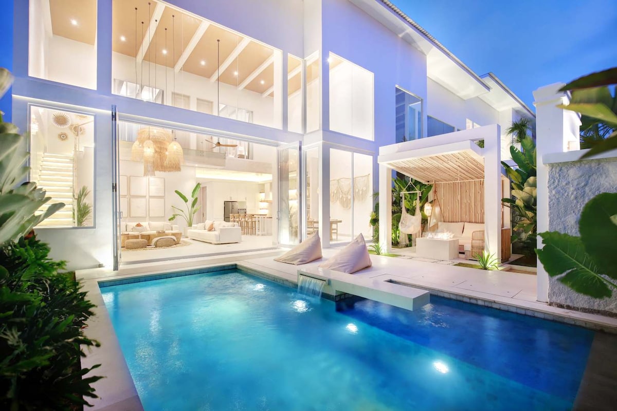 The outdoor area features a private pool with a calming blue hue, complemented by modern loungers and a cozy shaded cabana. The villa's expansive glass walls reflect the evening light, enhancing the connection between indoor and outdoor living spaces.