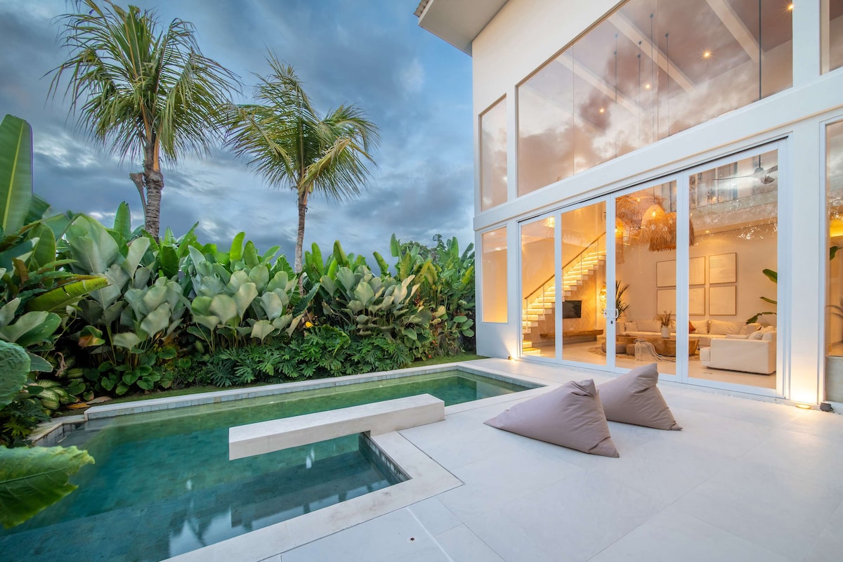 An outdoor area features a private pool surrounded by lush vegetation, providing a serene atmosphere. Large glass doors illuminate the space while leading to the interior. Cozy bean bags are positioned on the pool deck, offering a relaxed spot to unwind.