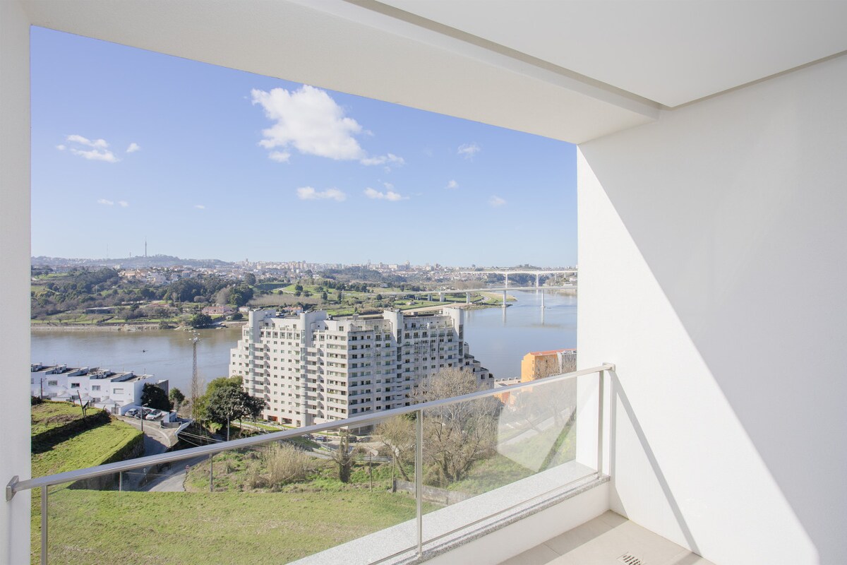 A spacious balcony features a railing and offers a panoramic view of the Douro River and surrounding landscape. Under a clear blue sky, the river reflects sunlight, while nearby buildings are visible along the riverbank.