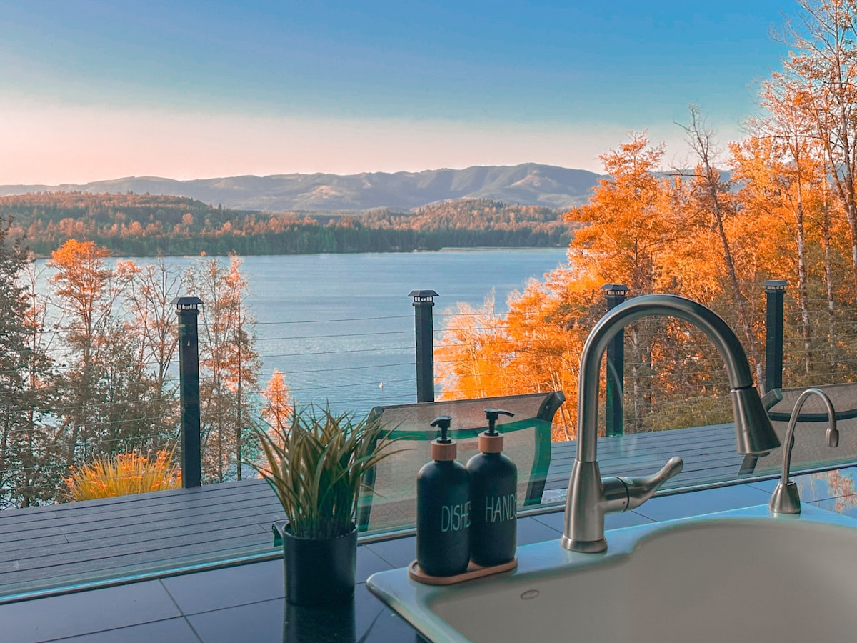 A view of Mayfield Lake is presented through large glass windows, highlighting vibrant autumn foliage along the shoreline. A modern kitchen sink is visible in the foreground, accompanied by a potted plant and decorative soap dispensers, enhancing the serene environment.