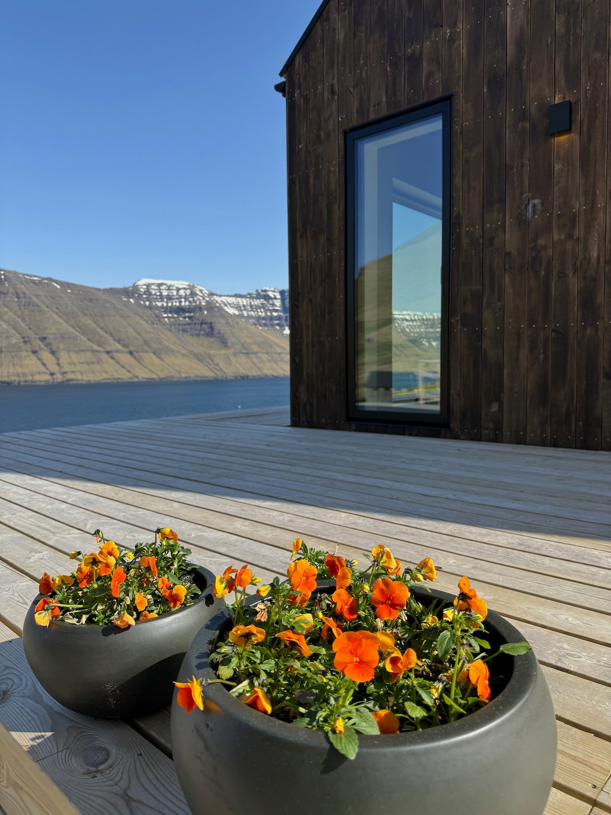 Two round planters filled with vibrant orange pansies are positioned on a wooden deck. A modern cottage with a large window reflects the surrounding landscape, showcasing a serene view of the water and mountains beyond.