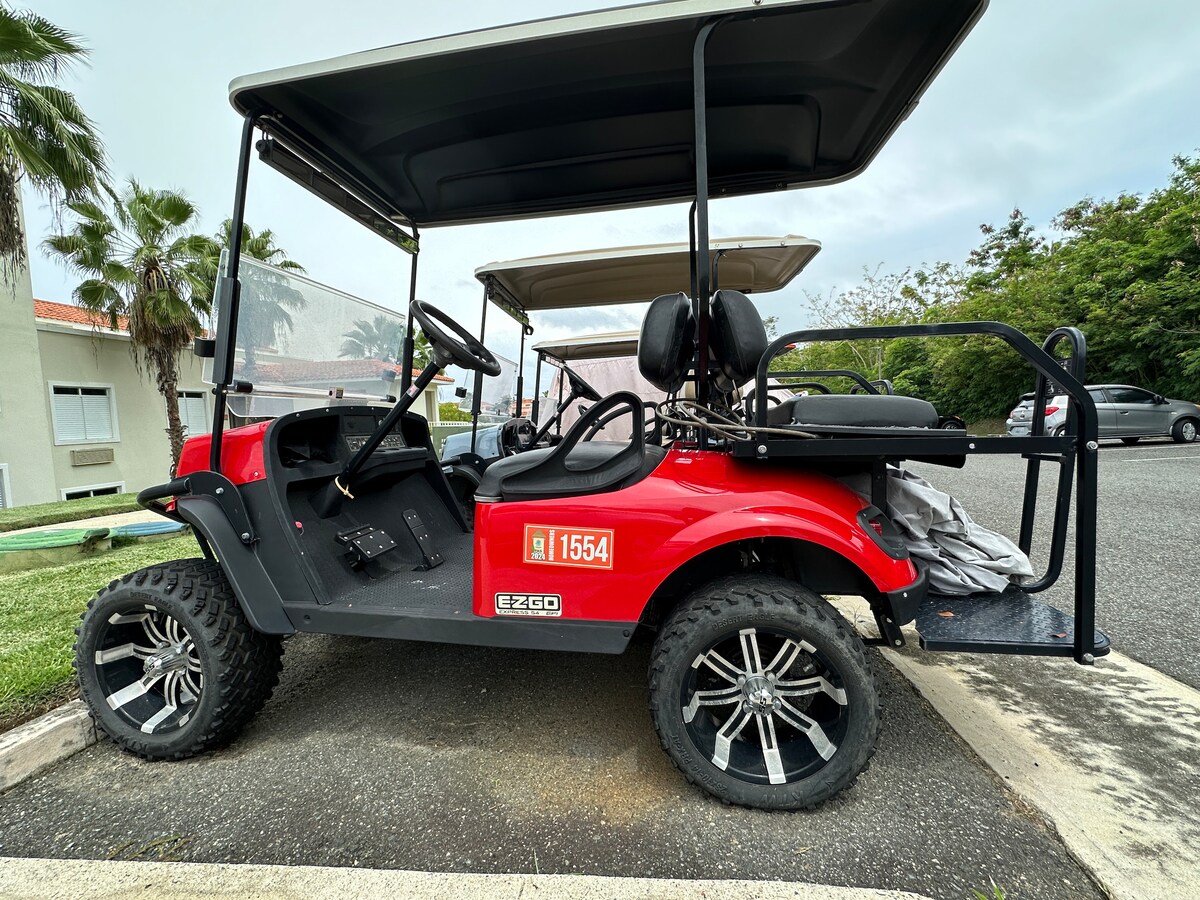 A red golf cart is parked on a paved area, featuring a black seating arrangement for two and spacious storage at the back. The vehicle showcases stylish wheels and is surrounded by greenery, adding a touch of vibrancy to the setting.