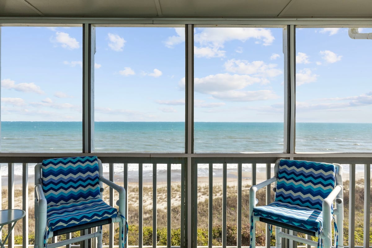 A screened-in balcony offers unobstructed views of the ocean, where waves gently meet the shore. Two chairs with a blue zigzag pattern are positioned for relaxation, complemented by a small side table. The scene is framed by expansive windows that allow ample natural light.