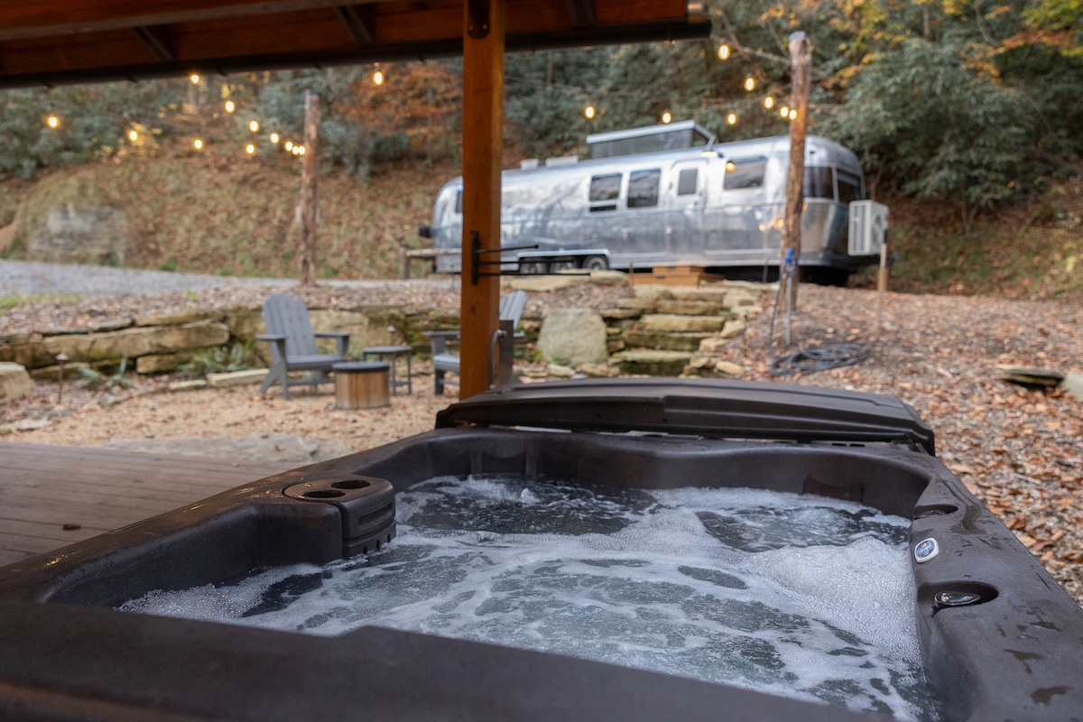 A hot tub is shown bubbling, surrounded by wooden decking and Adirondack chairs. String lights hang above, providing soft illumination. In the background, the shimmering exterior of an Airstream is partially visible, nestled among trees with fall foliage.