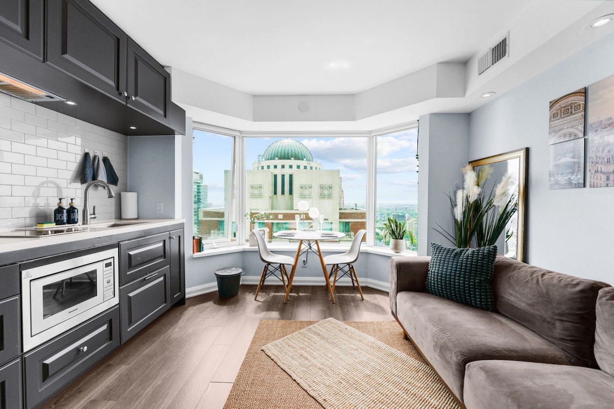 The living area features a modern kitchen equipped with appliances, including a microwave. A comfortable sofa is positioned adjacent to a bright window with a view of the Yorkville neighbourhood. A dining table with two chairs is set near the window, surrounded by plants.