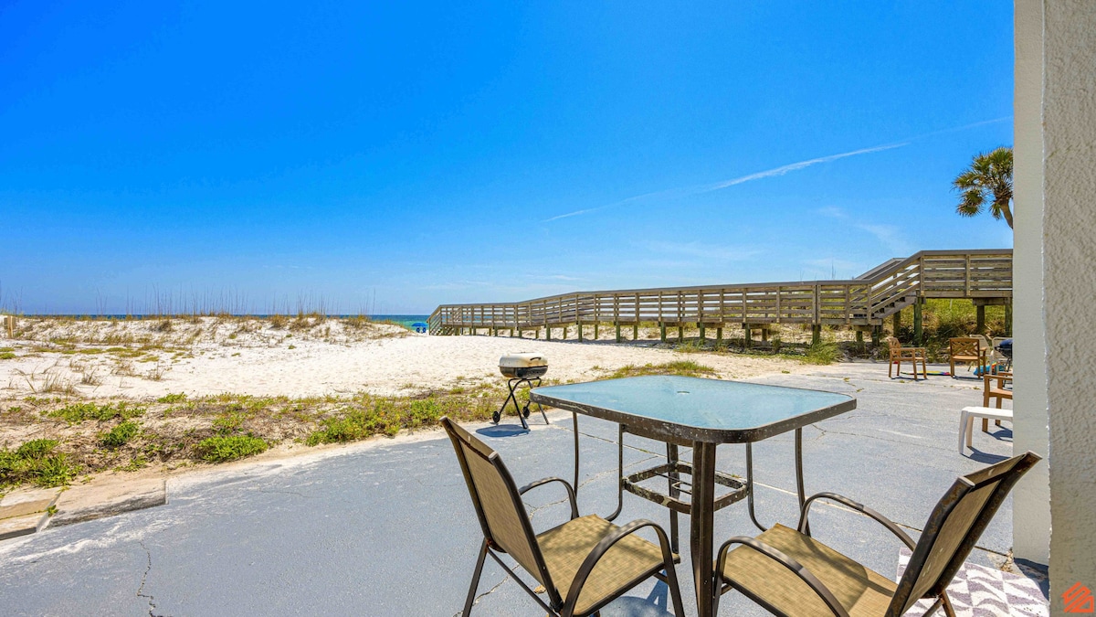 A private patio is visible, featuring a glass-top table and four metal chairs. Beyond the patio, a wooden boardwalk leads directly to the beach, bordered by soft white sand and coastal vegetation. The sky is clear with a bright blue hue, enhancing the beachfront view.