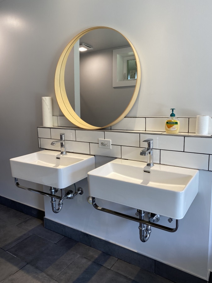 Two white wall-mounted sinks are positioned side by side under a round wooden-framed mirror. A bar of soap and a roll of paper towels are placed nearby on a tiled backsplash. Soft lighting illuminates the space, highlighting the clean lines and modern design.