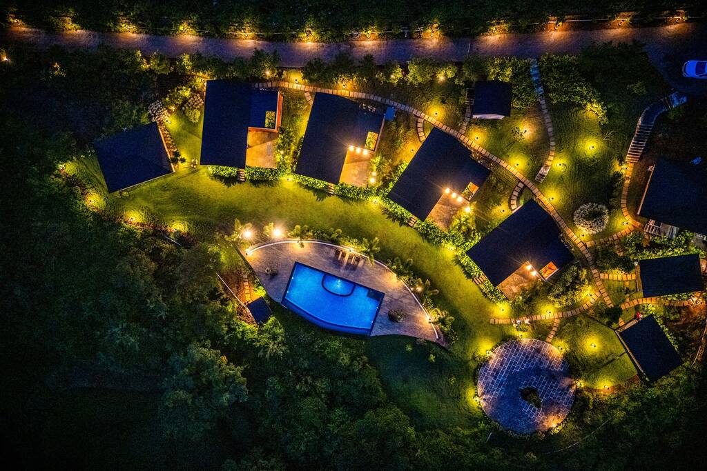 An aerial view of the property showcases several cabins arranged around a central swimming pool. Soft lights illuminate the pathways and gardens, creating a serene ambiance. The lush greenery and laid-out lawn complement the inviting pool area, enhancing the peaceful outdoor retreat.