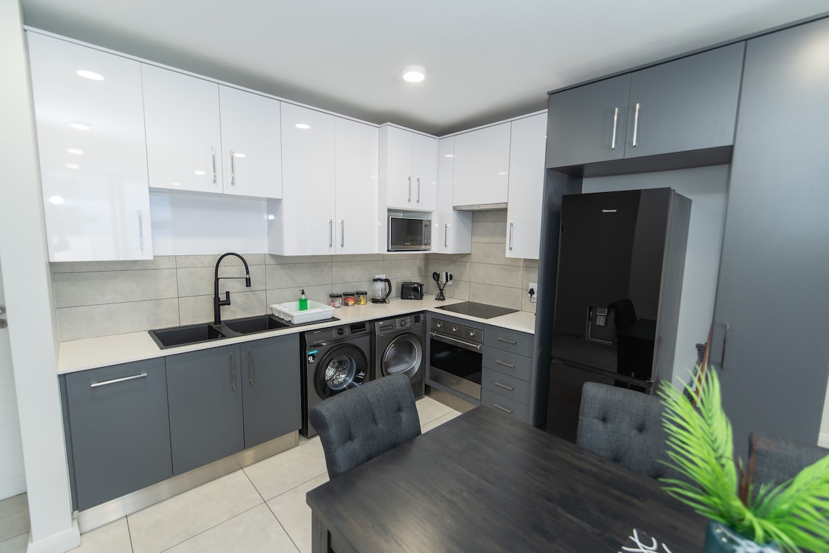 A modern kitchen is displayed featuring sleek cabinetry in white and dark grey. Stainless steel appliances, including a microwave and washer-dryer combo, are visible. A dark wooden dining table with tufted chairs is positioned nearby, accompanied by a decorative green plant for a touch of color.
