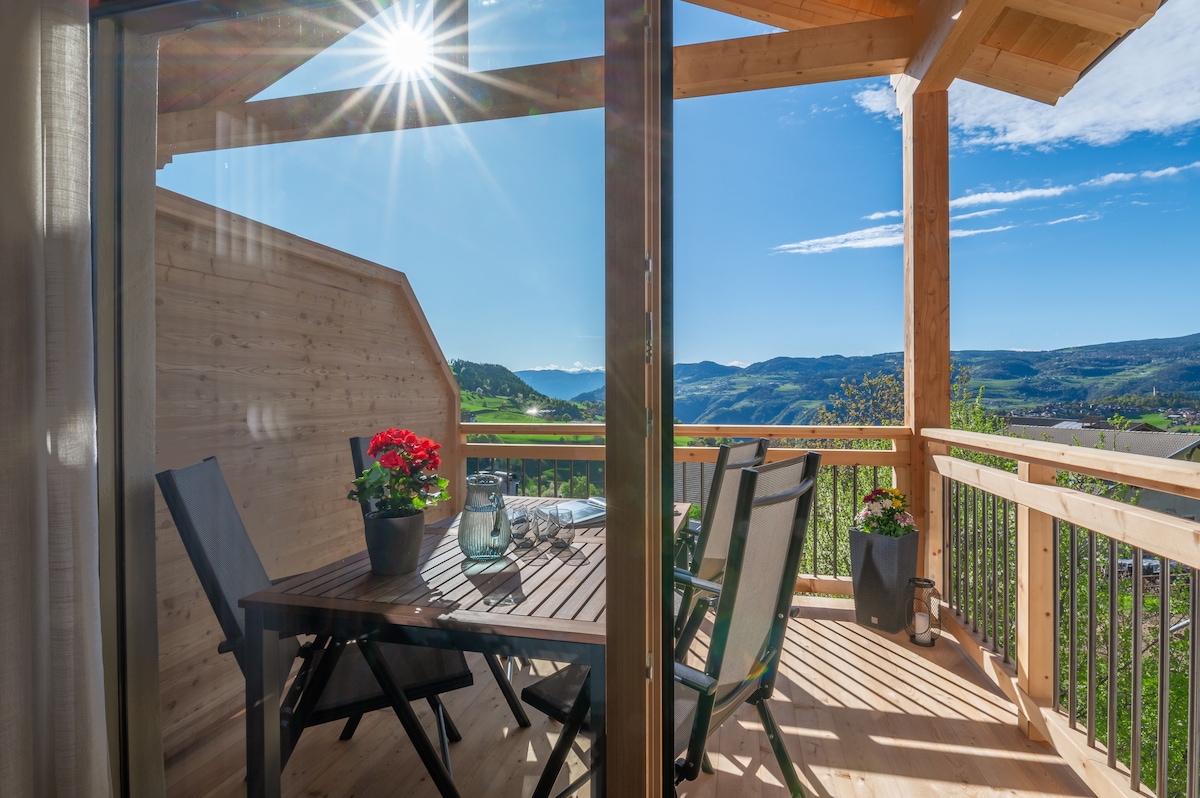 A private balcony is shown, featuring a wooden table surrounded by dark outdoor chairs. Potted flowers add a touch of color, while the expansive landscape of rolling hills and blue sky can be seen in the background. Sunlight filters through, creating a bright atmosphere.