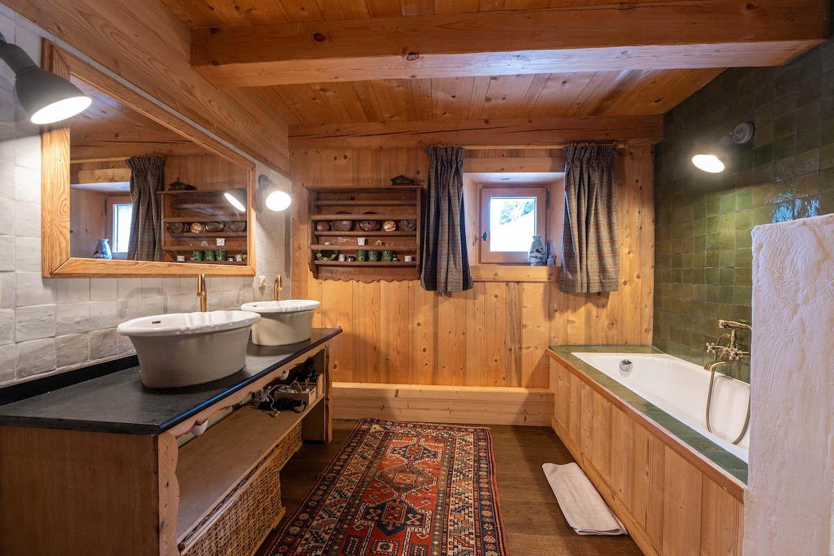 The spacious bathroom features double sinks set on a wooden vanity, with a large mirror above. A bathtub lies adjacent, complemented by natural wood finishes and decorative shelving. Warm lighting and window views enhance the calming atmosphere, while an area rug adds a touch of comfort.