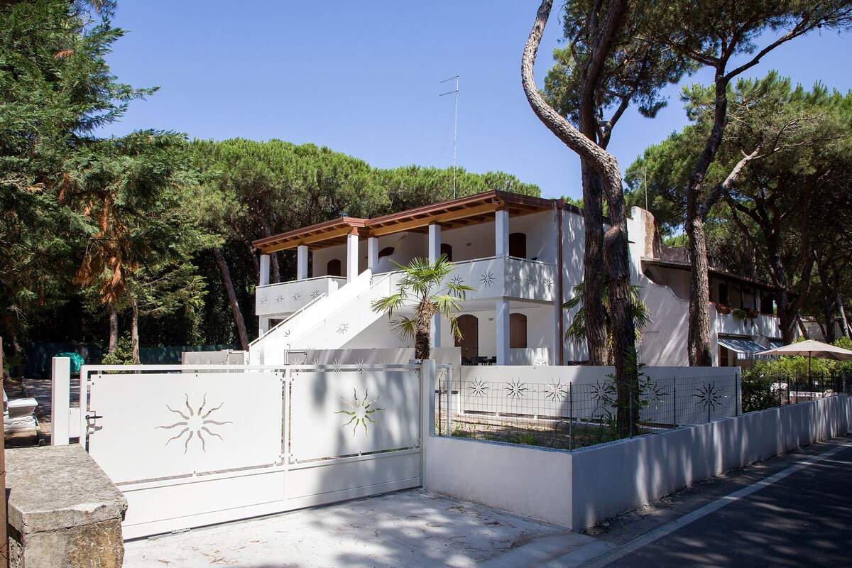 A two-story white building is situated amid tall pine trees, featuring a gated entrance with decorative elements. A parking area is visible alongside the paved road, with greenery surrounding the property. Sunlight reflects off the building's surfaces, enhancing the natural setting.