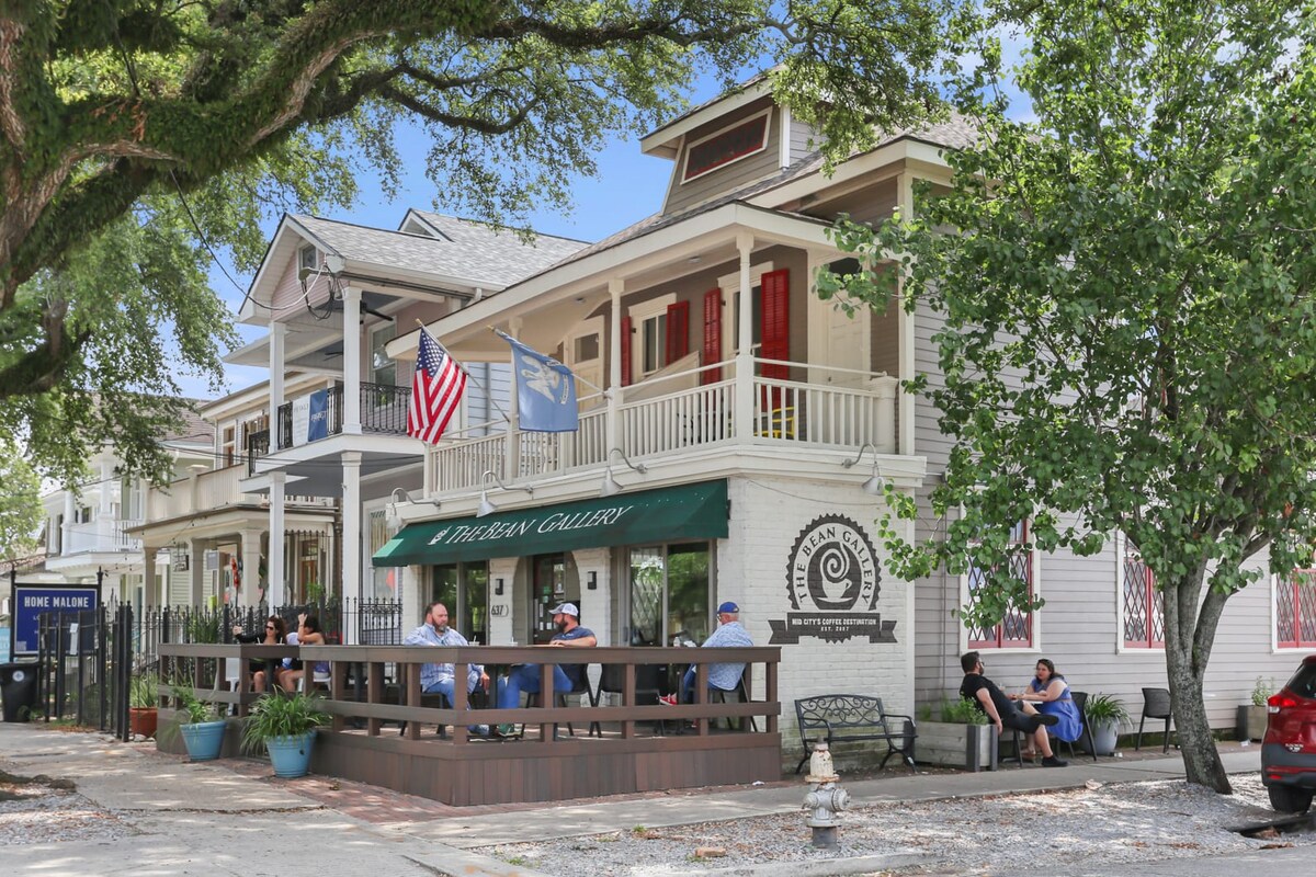 A row of charming two-story buildings is pictured, featuring balconies with white railings and vibrant red shutters. A covered outdoor seating area in front is occupied by guests enjoying drinks. The façade displays local art and flags, surrounded by lush greenery from nearby trees.