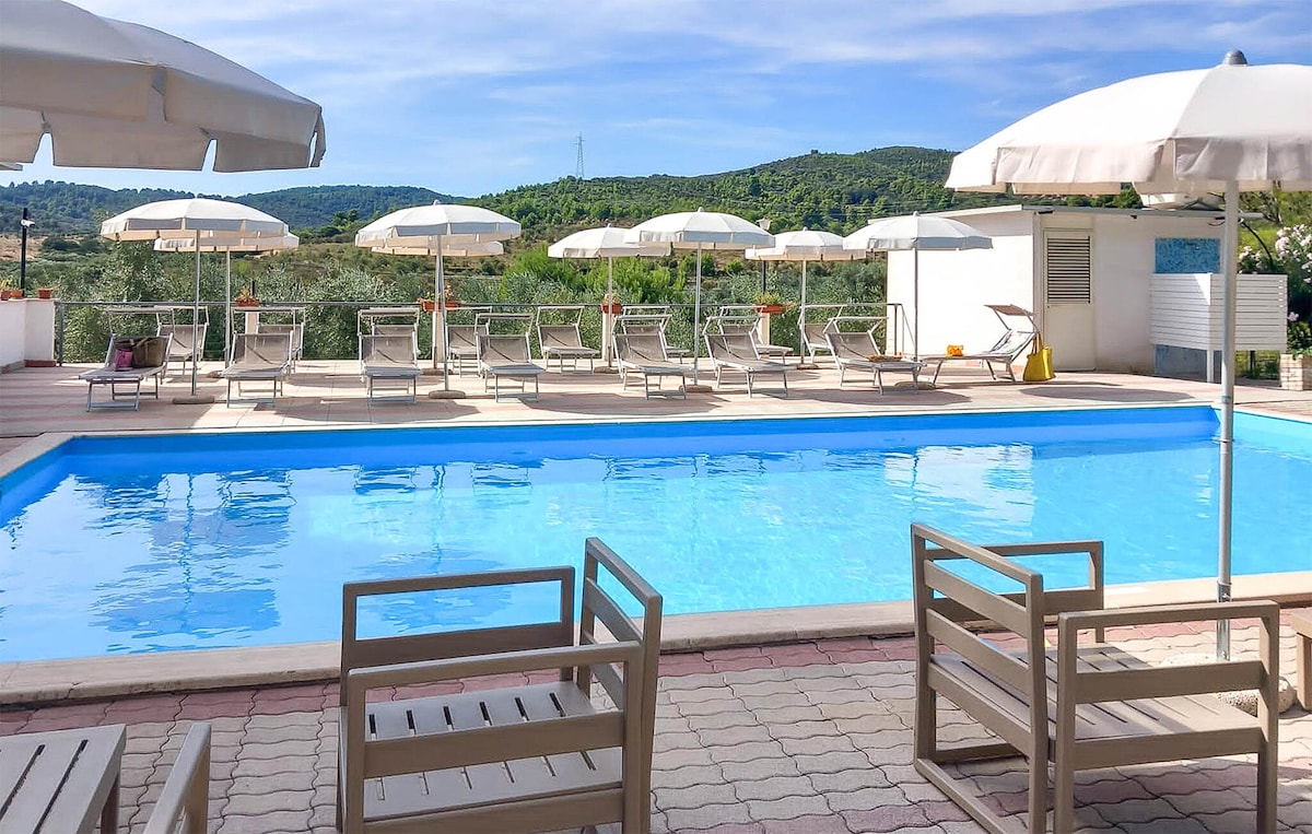 A shimmering swimming pool is surrounded by sun loungers and umbrellas, providing ample space for relaxation. The scenic view of rolling green hills is visible in the background, enhancing the tranquil outdoor setting.