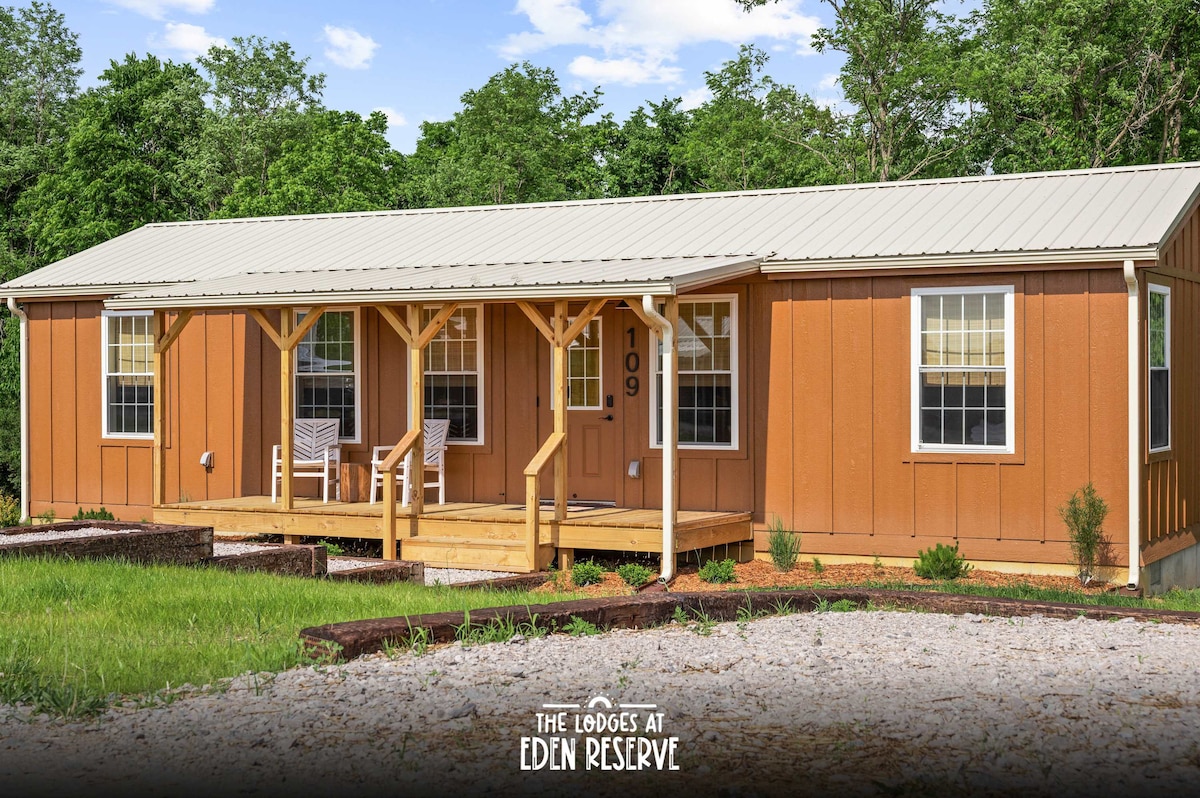 A spacious lodge exterior is shown, featuring a wooden porch with two rocking chairs. The building is painted in warm orange tones, complemented by white window frames. Surrounding greenery and a gravel pathway enhance the rural setting.