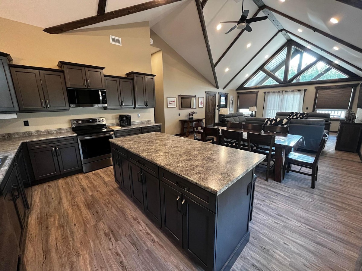 An open-concept kitchen features dark cabinetry and a large island with a textured countertop. The space includes stainless steel appliances and a dining area with ample seating. Tall ceilings with exposed beams add to the spacious feel, while natural light filters through the windows.