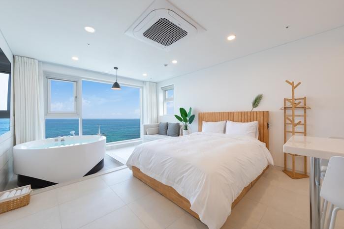An airy room features a spacious bed with crisp white linens and a cozy seating area by a large window. A bathtub with a view of the sea complements the relaxing atmosphere, while a small dining table and decorative plant add to the room's charm.