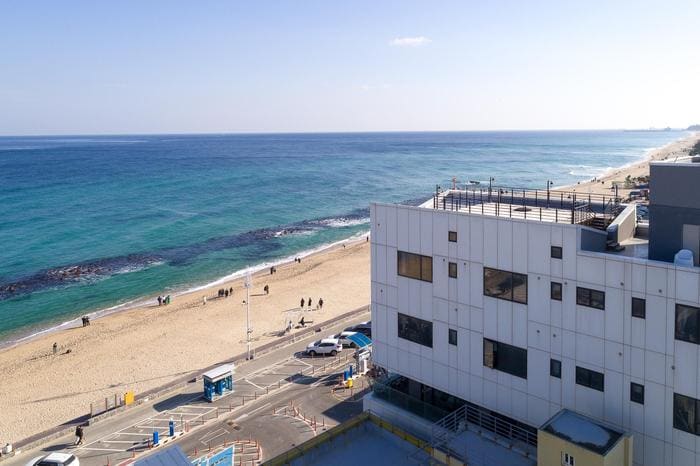 The image captures a view of a sandy beach and the ocean, with gentle waves lapping at the shore. A multi-story building is visible, showcasing large windows. People can be seen walking along the beach, emphasizing the coastal setting.