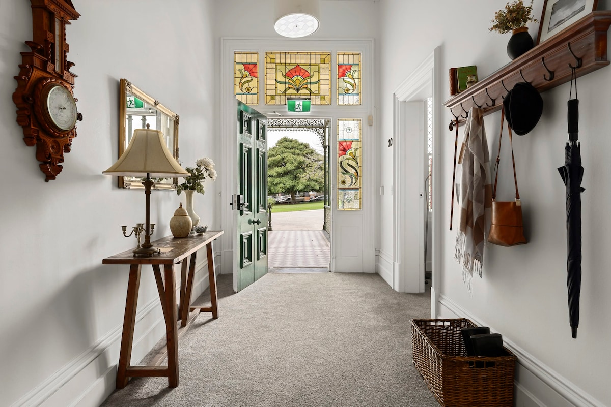 A bright entrance hall features a decorative stained-glass window above a prominent door. A small wooden table with a lamp is positioned against the wall. To the side, a coat rack holds various items, and a wicker basket sits on the carpeted floor.