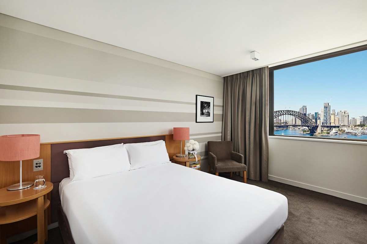 A welcoming hotel room features a spacious king-size bed dressed in crisp white linens. A side table with a lamp and a decorative vase is positioned next to the bed. A comfortable armchair sits nearby, and large windows offer views of the Harbour Bridge and city skyline.