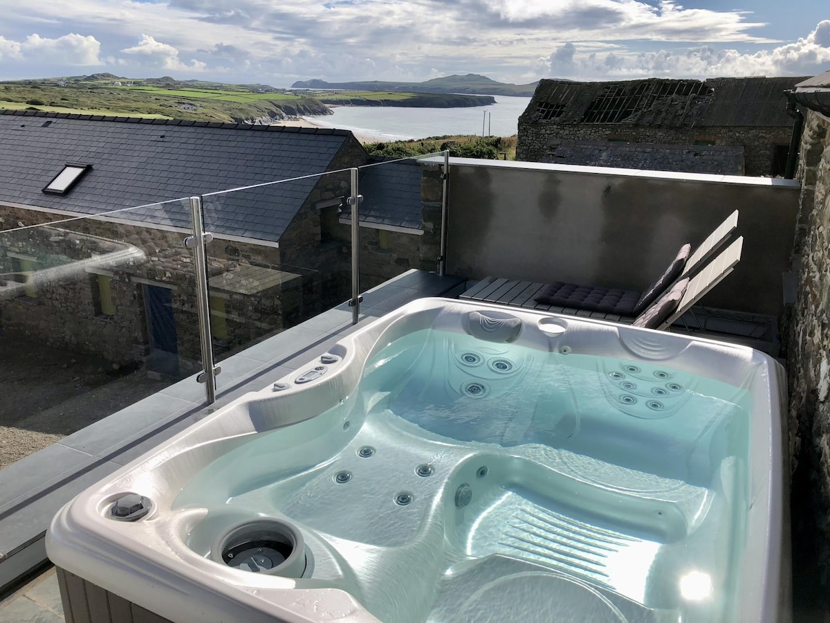 A spacious hot tub is situated on a newly built stone veranda, with clear views of the coastline and distant hills. Lounge chairs are positioned nearby, inviting relaxation in the outdoor space. The serene atmosphere is enhanced by the surrounding landscape.