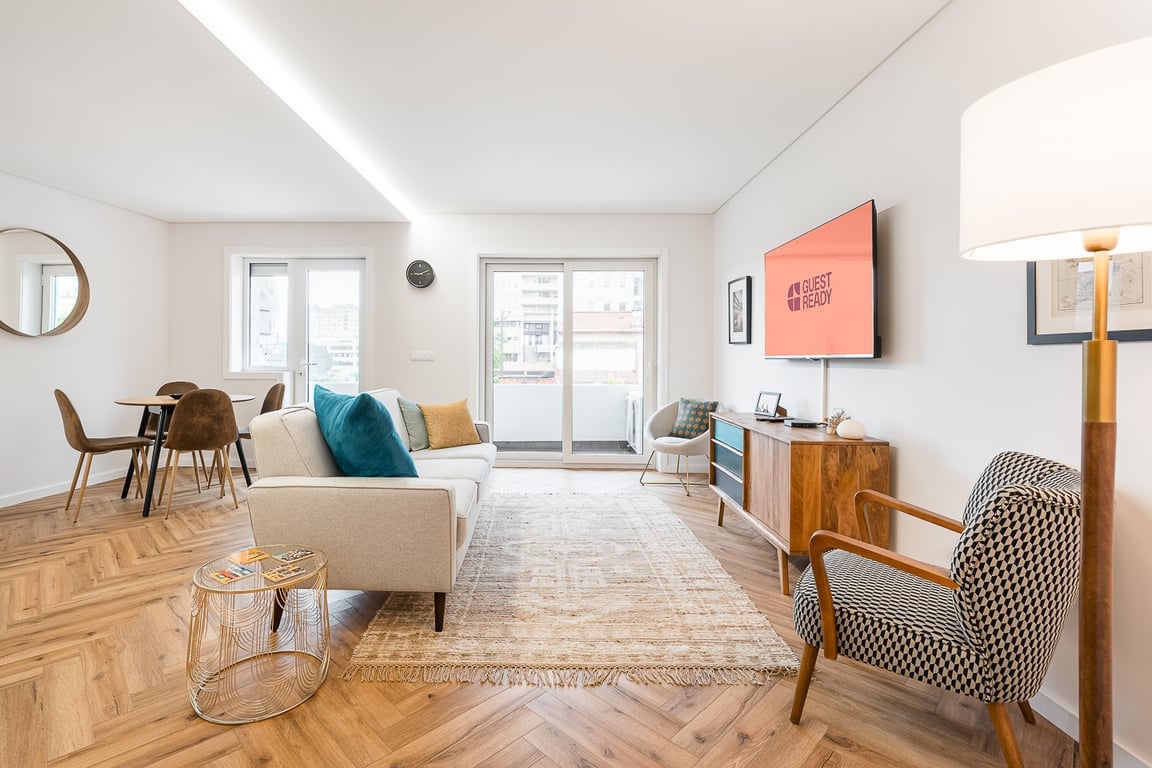 The open living area features a comfortable sofa, stylish accent chairs, and a wooden media console with a television. Natural light floods the space through large windows, with a dining table set for four in the background. A patterned rug adds texture to the warm wood flooring.