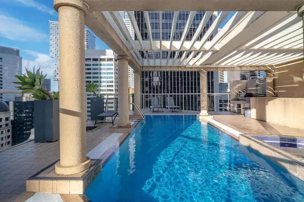 A rooftop pool area is showcased with a clear blue water surface beneath a partially shaded deck. Vertical columns support a pergola overhead, contributing to an open ambiance. Surrounding the pool, lounge chairs are positioned along the edge, with city skyline views visible in the background.