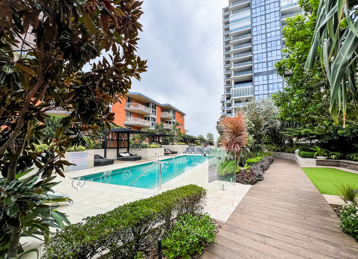 A landscaped outdoor area surrounds a modern pool, showcasing clear water and comfortable lounge chairs. The pathway is lined with greenery and vibrant plants. Nearby buildings and a gazebo are visible, creating a serene environment for relaxation.
