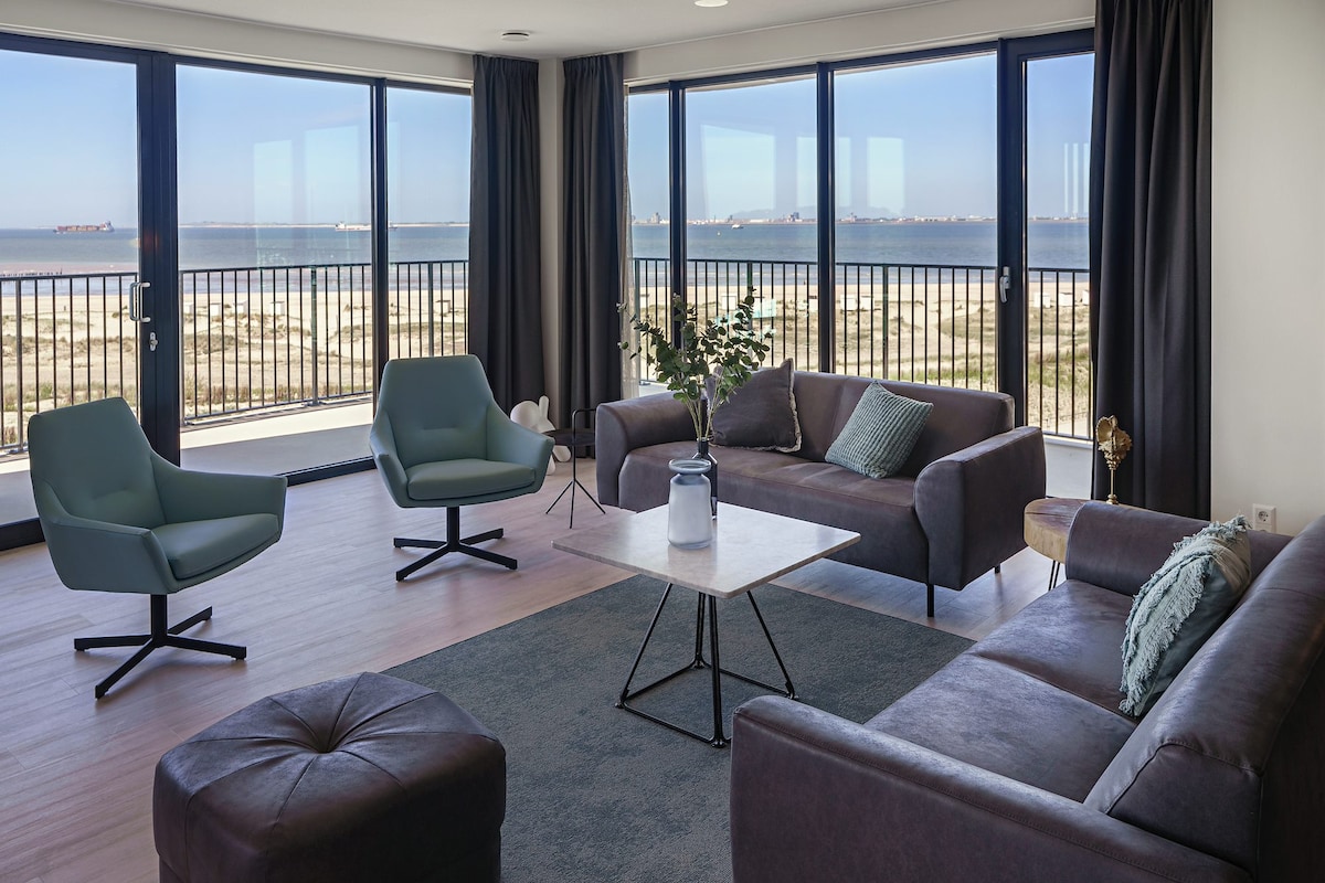 The spacious living room features large glass doors that offer stunning views of the North Sea. Two contemporary armchairs and a sofa provide ample seating, complemented by a coffee table at the center. Natural light fills the room, enhancing the open and airy feel.